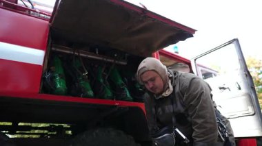 Genç itfaiyeci büyük kırmızı bir arabanın arka planına kask ve kar maskesi takıyor. İtfaiye arabasının yanında tam teçhizatlı erkek itfaiyeci çalışmaya hazır. Hayat kurtarma kavramı ve kahramanlık mesleği.