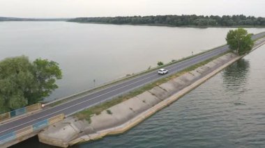 Beyaz arabanın havadan çekilmiş görüntüleri yaz günü baraj yolundan geçer. SUV göl ya da göl kenarındaki yolda gidiyor. Oto üzerinden uçmak nehir köprüsünden geçer. Hafta sonu seyahati kavramı. Üst görünüm