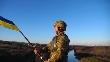 Tepede dikilen askeri üniformalı asker Ukrayna bayrağını kaldırdı. Elinde bayrak sallayan Ukraynalı asker. Rus saldırısına karşı zafer. İstila direnci konsepti. Yavaş çekim.