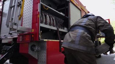 Tam teçhizatlı erkek itfaiyeci büyük kırmızı kamyonun yanındaki yangın hortumunu çözüyor. Profesyonel itfaiyeci yangını söndürmeye hazırlanıyor. Kahramanlık mesleği ve hayat kurtarma kavramı. Yavaş çekim.
