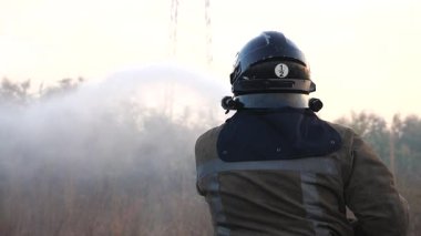 Tam teçhizatlı profesyonel itfaiyeci sahada alev saçıyor. Üniformalı erkek itfaiyeci, kırsal bölgelerde yangın hortumuyla yanan kuru otları söndürüyor. Kahramanlık mesleği ve yangın güvenliği kavramı.