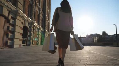 Şık bir kadının arka görünüşü, şehir caddesinde yürüyen kağıt paketler taşıyor. Zarif genç kız alışveriş sonrası kaldırıma çıkan alışveriş torbalarını tutuyor. Mevsimlik satış ve indirim kavramı. Yavaş çekim.