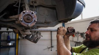 Üniformalı otomobil tamircisi, iş yerinde ingiliz anahtarı kullanarak arabanın tamirini yapıyor. Profesyonel erkek tamirci garajdaki bir kaldırma aracının altında çalışıyor. Otomobil bakımı kavramı.