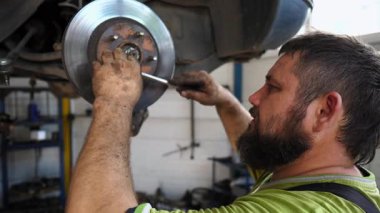 Garajda tornavida kullanan profesyonel sakallı otomobil tamircisi. Yetişkin erkek tamirci atölyede kaldırma aracının altında çalışıyor. Otomobil bakım kavramı.