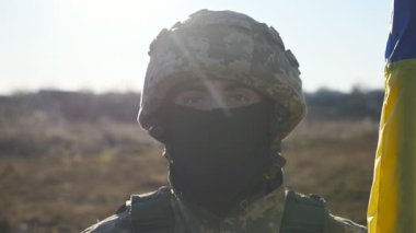 Miğferli ve kar maskeli Ukraynalı erkek asker güneş ışığının arka planına karşı milli bayrak sallıyor. Kırsal kesimde Ukrayna bayrağı taşıyan genç bir askerin portresi. İstila direnişi.