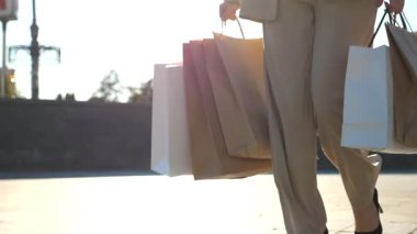 Hands of young woman carry shopping bags walking along urban street. Female arms hold paper packages going on city square after purchases. Concept of sales and discounts. Slow motion Close up.