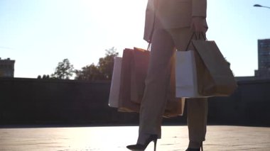 Young beautiful woman holds shopping bags going at urban street after purchases. Fashionable lady carries paper packets walking along city square. Concept of leisure, sales and discounts. Slow motion.