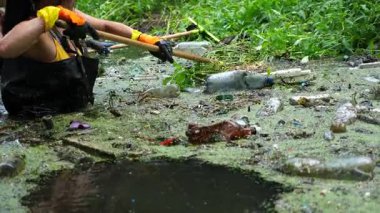 Koruyucu eldivenli kadın çevrecilerin elleri enkaz nehirlerini temizliyor. Kirli gölde plastik şişe toplayan kauçuk elbiseli tanınmayan gönüllüler. Çevre kirliliği koruması.