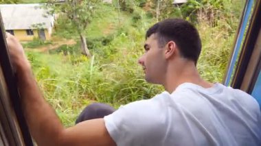 Hareket halindeki trenin kapılarında oturan ve güzel doğayı kollayan mutlu adamın yan görüntüsü. Yolcu trenine binen ve yaz yolculuğunun tadını çıkaran genç bir adam. Kapat..