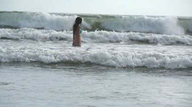 Bikinili genç bir kız okyanusta onu kaplayan dalgaların arasında duruyor. Mayolu güzel bir kadın denizde rahatlar ve yolculuktan zevk alır. Yaz tatili ya da tatil kavramı. Yavaş çekim.