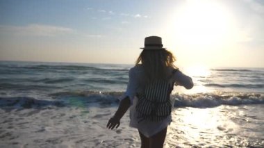 Bikinili, sırt çantalı, denize koşan ve ayaklarıyla su fışkırtan mutlu genç kız. Güzel bir kadın sahilde dönüyor ve dışarıda eğleniyor. Yaz tatili kavramı. Yavaş çekim.