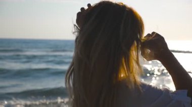 Bikinili ve tişörtlü genç bir kız kıyıda yürüyor ve sarı saçlarıyla oynuyor. Güneş gözlüklü güzel kadın yaz tatilinin tadını çıkarıyor. Arka planda deniz manzarası. Yavaşça kapat..