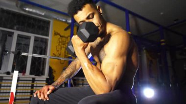 Kaslı Afro-Amerikan sporcu, spor kulübünde pazılarını dambılla eğitiyor. Modern spor salonunda kaslarını çalıştıran Afrikalı bir sporcu. Spor salonunda egzersiz yapan atletik siyahi adam. Sağlıklı yaşam tarzı.