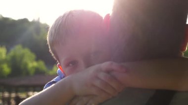 Kırsal kesimde küçük oğlunun elini tutan genç bir baba. Küçük çocuk kameraya bakıyor ve arka planda gün ışığına karşı babasına sarılıyor. Aile mutluluğu kavramı. Yavaşça kapat..