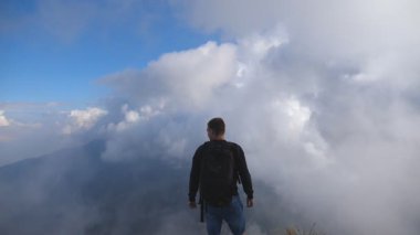 Sırt çantalı genç bir yürüyüşçü dağın kenarında duruyor. Arka planda bulutlu bir gökyüzü ve seyahat sırasında özgürlüğün tadını çıkarıyor. Güzel manzara. Yavaş çekim.