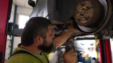 Üniformalı profesyonel sakallı tamirci atölyede çekiç kullanan arabanın tamirini yapıyor. Yetişkin erkek tamirci garajdaki bir kaldırma aracının altında çalışıyor. Otomobil bakımı kavramı.