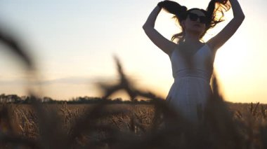 Buğday tarlalarının arasında duran çekici kadın günbatımı arka planında saçlarıyla nazikçe oynuyor. Genç kız akşam vakti dışarıda eğleniyor. Özgürlük kavramı. Doğayla uyum. Yavaş çekim.