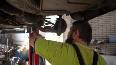 Erkek oto tamircisi atölyede kaldırma aracının altında çalışıyor. Yetişkin profesyonel tamirci serviste ya da garajda araba tamir ediyor. Düzenli otomobil bakımı kavramı. Yavaşça kapat..