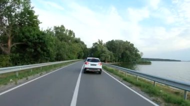 İnsansız hava aracı baraj yolundan geçen beyaz arabayı takip ediyor. SUV nehir ya da göl kenarındaki güzel kırsal yolda gidiyor. Yolculuk ya da doğa yolculuğu. Tatil aile seyahati kavramı. Havadan çekim Ağır çekim
