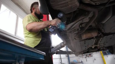 Skilled automotive technician working under a car in a garage, using a power tool to repair components, showcasing the intricate process of vehicle maintenance and repair in a professional setting.