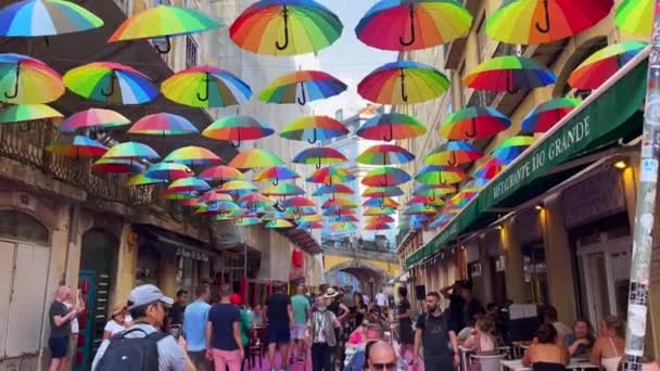 リスボンの建物の間にカラフルな虹色の傘がかかっているピンクの通りにぶら下がっている観光客 — ストック動画