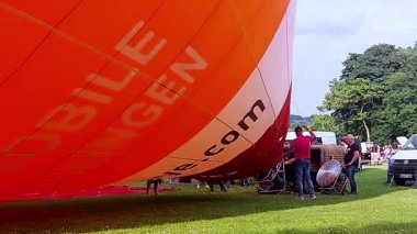 Almanya, Bonn 09.06.2025. Canlı turuncu bir sıcak hava balonu bir festivalde şişiriliyor. Erkekler ve kadınlar da dahil olmak üzere çeşitli yetişkinler, çimenli bir alanda hazırlanmaya yardımcı olurlar..