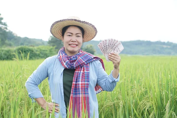 Asian farmer woman is at paddy field, hold Thai banknote money. Concept : Farmer happy to get profit, income, agriculture supporting money. Proud on crops.