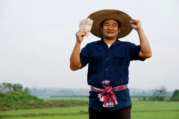 Happy Asian man farmer is at paddy field, holds Thai banknote money.  Concept : Happy farmer get profit, income, agriculture supporting money. Proud on crops.