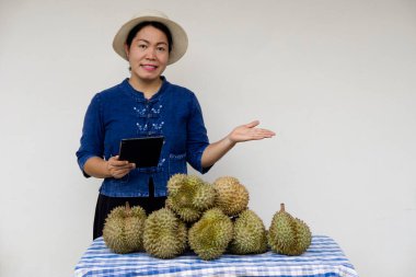 Asyalı kadın bahçıvan şapka takıyor, akıllı tablet tutuyor, Durian meyvelerinin yanında duruyor. Meyve satıcısı internet üzerinden tarım ürünleri satmak için internet teknolojisi kullanıyor. Satmak için durian yetiştir.