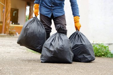 Closeup man holds black plastic bag that contains garbage inside, stand in front of house. Concept : Waste management. Environment problems. Daily chores. Throw away rubbish .             