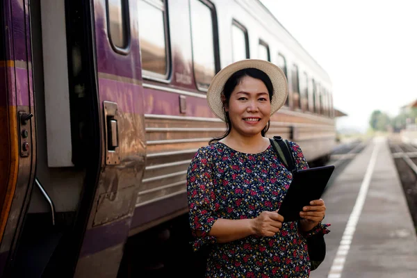 Happy Asian middle aged woman traveller is at railway station, wears hat, holds smart tablet and smiles. Concept, travel by train in Thailand can book or by ticket online.
