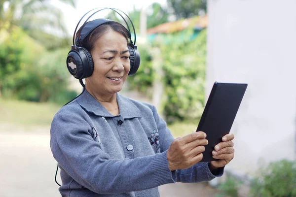 Happy Asian senior woman wears headphones, holds smart tablet, listen to music. Concept, elderly use technology smart device. Relax with online music, watch vedio. Learning new thing from internet.