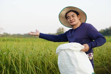 Handsome Asian man farmer is at paddy field, wears hat, holds white sack of organic fertilizer, feels confident. Concept, farmer satisfied in product for agriculture.                        