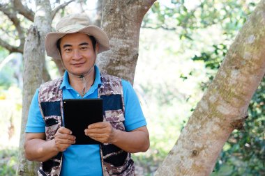 Asian man botanist is in forest, hold smart tablet to survey and collect information of botanical plants by using smart tablet. Concept , adventure, field research about environment.      