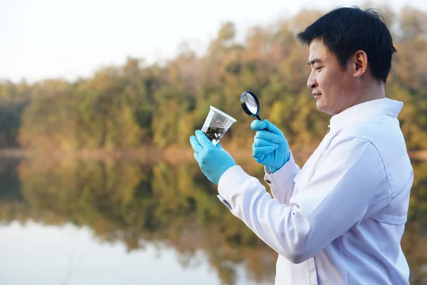 Asian man ecologist holds magnifying glass to inspect water in transparent glass at the lake. Concept, explore, analysis water quality and creature from natural source. Ecology field research.