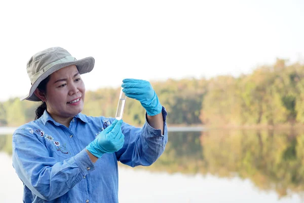 Asian woman environment researcher holds tube of sample water to inspect from the lake. Concept, explore, analysis water quality from natural source. Ecology field research.