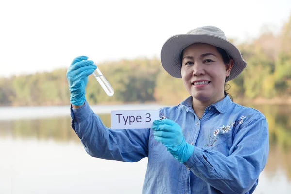 Asian woman ecologist hold tube of sample water and tag with word Type 3 for inspect and research water quality at the lake. Concept, explore, analysis water quality from natural source. Ecology field