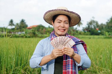 Asian farmer woman is at paddy field, wears hat and red plaid shirt, hold Thai banknote money.  Concept : Farmer happy to get profit, income, agriculture supporting money.   