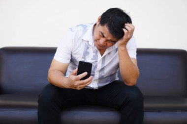 Asian man sits on sofa, hold smartphone, feels headache. Upset, sad. Concept, know bad news from social media online. Feeling and emotion expression. drepression, failure, disappointment, broken heart