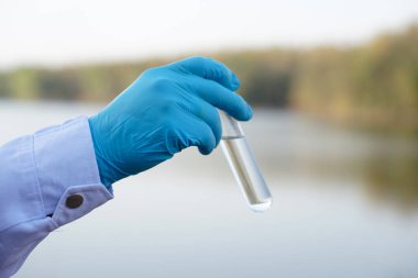 Closeup researcher hands wears blue gloves holds test glass tube that contain sample water from the lake. Concept, explore, inspect quality of water from natural source.           
