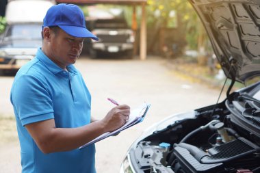 Asyalı tamirci mavi kep ve mavi gömlek giyer, elinde kağıt tutar, arabanın motorunu kontrol eder ve analiz eder. Konsept, açık hava araba teftiş servisi. Kaza sigortası talep et.. 