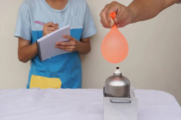 Balloon with water filled is burned on fire flame of metal lantern. Student observe and take note on paper notebook. Concept, science experiment. Fireproof balloon demonstration. Learning by doing.