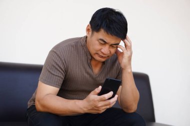 Asyalı adam kanepeye oturur, telefonu tutar, baş ağrısını hisseder. Üzgün, üzgün. Çevrimiçi sosyal medyadan kötü haberleri bilmek kavramı.      