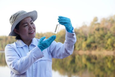 Asian woman environment researcher holds tube of sample water to inspect from the lake. Concept, explore, analysis water quality from natural source. Ecology field research.      