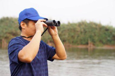 Asyalı adam mavi başlık takıyor, dürbünü gölde tutuyor, doğa kaynağı. Kavram, doğa keşfi. Ekoloji dersi. Hobi aktivitesi, yaşam tarzı. İnsan ortamı keşfeder  