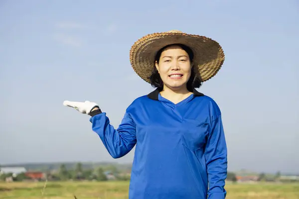 stock image Portrait of Asian woman farmer wears hat and blue shirt, make gesture to present product for advertisement. Concept : Agriculture occupation. , Sky background.