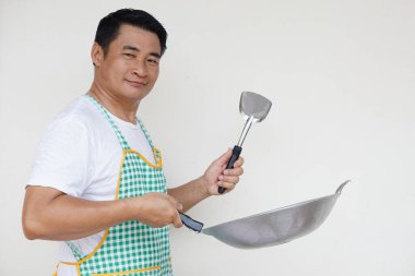 Yakışıklı Asyalı adam yemek pişiriyor, beyaz gömlek ve önlük giyiyor, elinde kızartma tavası ve kepçe spatula tutuyor. Kavram, yemek yapmayı seviyorum. Mutfak yaşam tarzı. Kameraya bak. Metin için alanı kopyala     