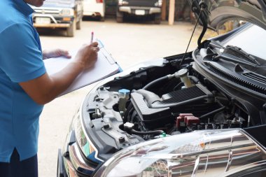 Yakından tamirci, arabanın motorunu inceliyor. Kağıt tutuyor, arabanın motorunu kontrol ediyor ve analiz ediyor. Konsept, açık hava araba teftiş servisi. Kaza sigortası talep et..   