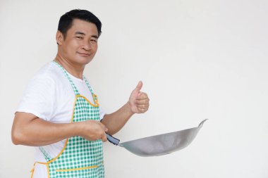 Yakışıklı Asyalı adam yemek pişiriyor, beyaz gömlek ve önlük giyiyor, kızartma tavası ve baş parmağını kaldırıyor. Kendine güven. Kavram, yemek yapmayı seviyorum. Mutfak yaşam tarzı. Kameraya bak. Metin için alanı kopyala 