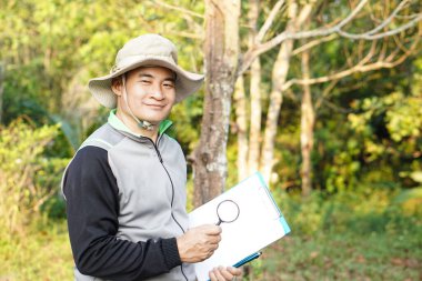 Asyalı bir botanik uzmanı kağıt not defteri tutar ve ormandaki bonik bitkileri incelemek için büyüteç kullanır. Kavram, araştırma, botanik bitkileri. Orman ve çevre koruma.    